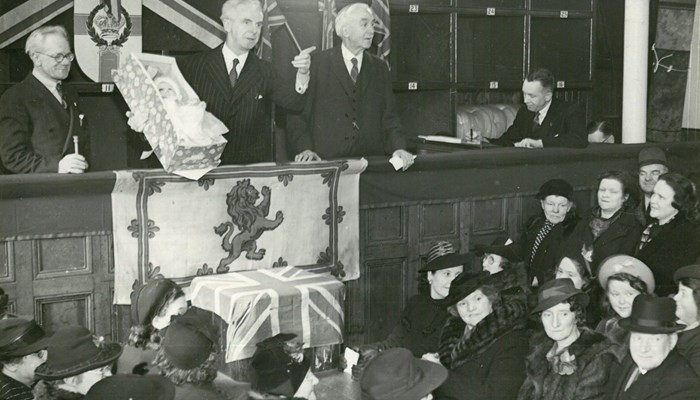 Three people standing talking to a crowd lower below, holding a baby in a small crib with a Lion Rampant of Scotland flag and a Scotland flag draped below.
