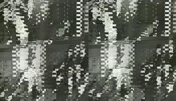 Three people standing talking to a crowd lower below, holding a baby in a small crib with a Lion Rampant of Scotland flag and a Scotland flag draped below.