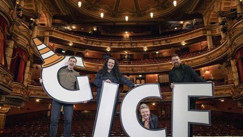 A group of four comedians are standing on a theatre stage with an empty auditorium behind them. They are each carrying a large cut-out letter to spell out GICF