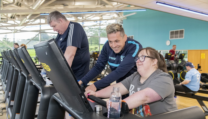 Three adults are smiling and exercising together on treadmills in a brightly lit gym.
