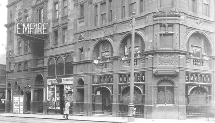 A black and white photo of a large tenement style sandstone building with lots of architectural designs and signs for a theatre on the outside.