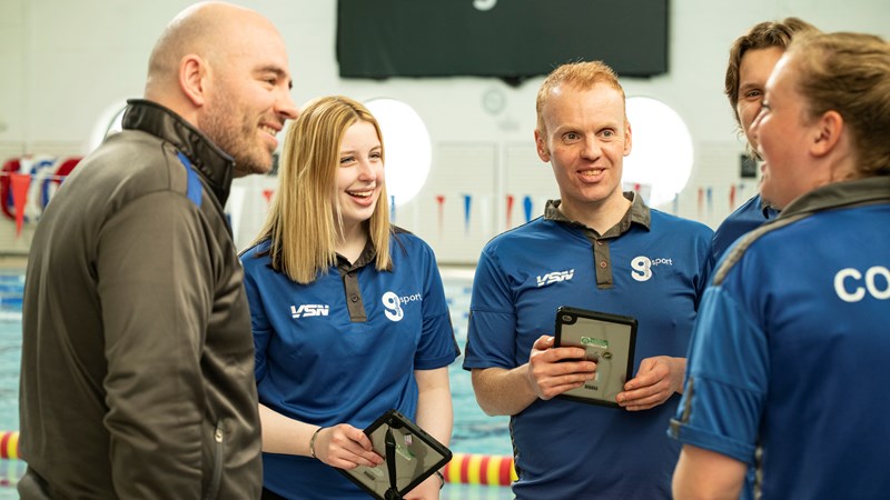 Five Glasgow Life staff smiling and chatting at a swimming pool venue. They are wearing Glasgow Life branded clothes and there is a branded black Glasgow Life sign in the background.