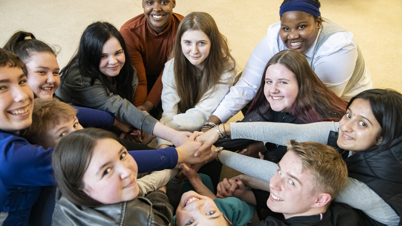 A group of young people sit close together in a circle, extending their arms toward the center where their hands are stacked on top of each other. The scene is indoors, with everyone leaning in from different directions, forming a tight, collaborative composition. The image captures multiple faces arranged around the edges of the frame, with overlapping hands at the centre, highlighting teamwork and unity.