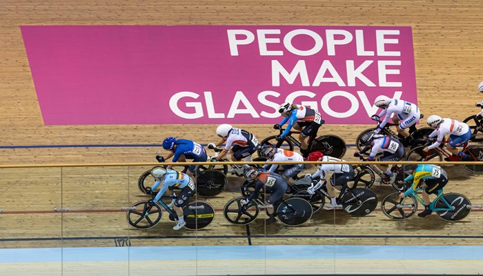 Cyclists on an indoor track ride past a pink and white sign which reads 'People Make Glasgow'