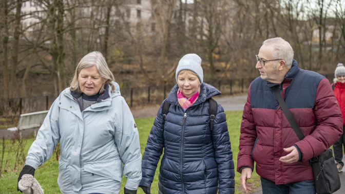 Three senior walkers in winter coats and hats talking while walking along a path in a park.