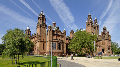 A red sandstone building beneath a blue sky is surrounded by green trees