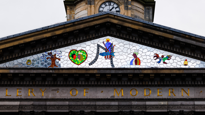 A neoclassical building’s pediment decorated with a colorful mosaic featuring symbolic figures, shapes, and animals sits above a stone façade with gold lettering. Above the pediment, a clock tower rises with a round clock face showing the time around 10:10.