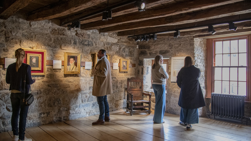 4 people are standing in a dimly lit room with stone walls and wooden beams across the ceiling. They are looking at framed portraits
