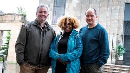 Three figures stand together in front of a brick building 