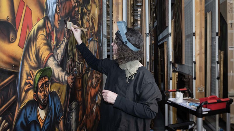 Conservator restores a large painting in a storage area with racks of framed artworks.