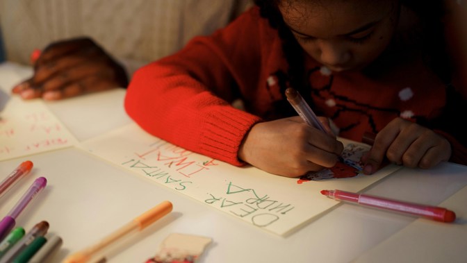 A young person drawing on a large sheet of white paper while leaning on a table, they are wearing a red jumper and using different coloured pens.