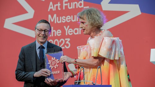 Two people on a stage at an award ceremony. One is holding a glass trophy with Winner written on it. Behind them is a large sign that reads Art Fund Museum of the Year 2023