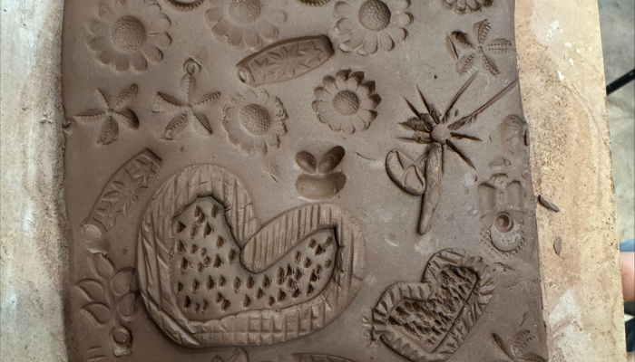 A square slab of brown clay held at the bottom corners by two hands. The surface is covered with stamped and carved patterns, including flowers, leaves, hearts, and other decorative shapes, creating a textured, layered design.