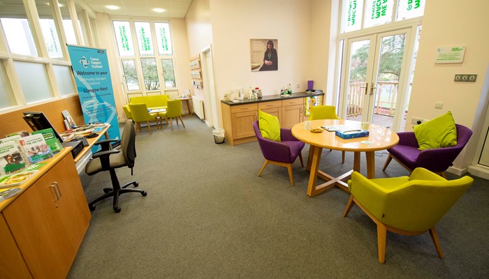 Health and Wellbeing area with 2 tables surrounded by soft purple and green chairs. There is also a desk with a PC at the side of the room. The room has large, tall windows and there are patio doors near one of the tables 