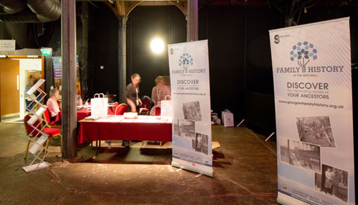 A photo of staff setting up a stall at an event with a red table cover, Family History pop up banners, leaflets and goody bags.