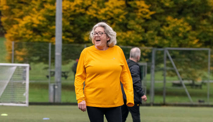 A smiling older person in a bright orange top walks towards an orange football on an outdoor pitch.