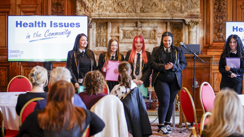 Five school students in uniform are giving a presentation to an audience in an ornate banqueting hall with marble fireplace. They have a microphone and projector screen