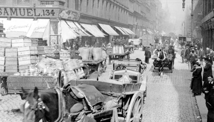 A photo of a busy Glasgow street filled with shoppers, tall buildings and horse drawn carriages stocked high with goods.