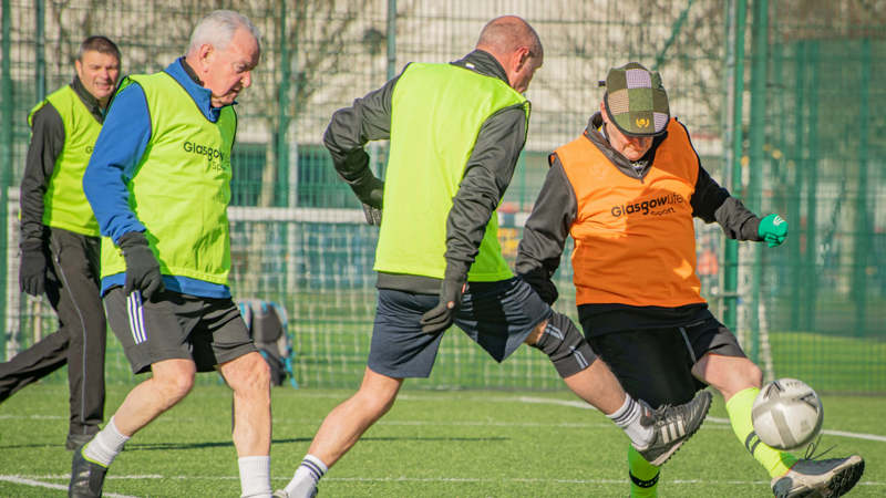 Four football players on an outdoor green pitch battle over the ball. The players are older adults and wear yellow and orange vests.