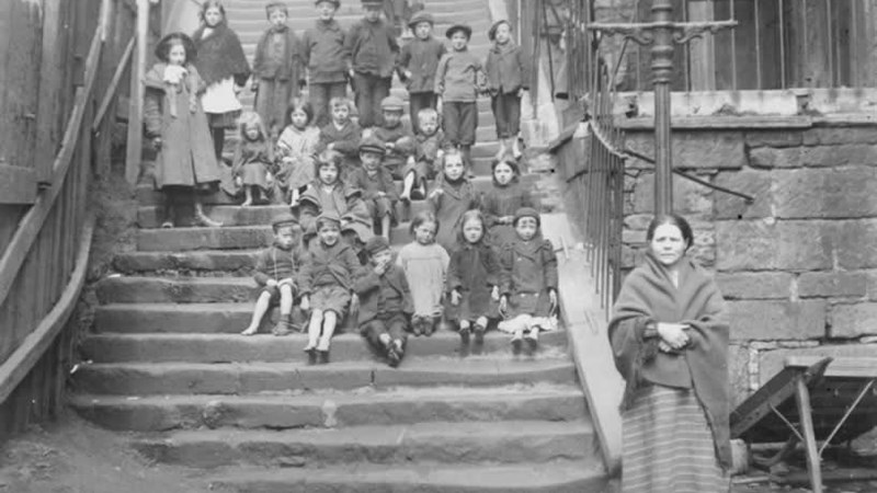 Black and white photo showing a women standing at the bottom of a set of stairs with a large group of children of various ages standing on the steps behind her