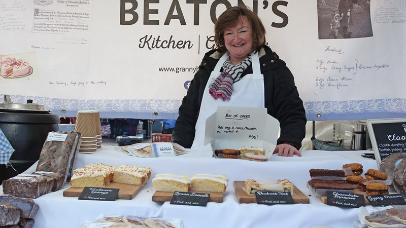 A woman stands behind her bakery stall, which displays a variety of baked good. A banner behind her reads "Granny Beaton's"