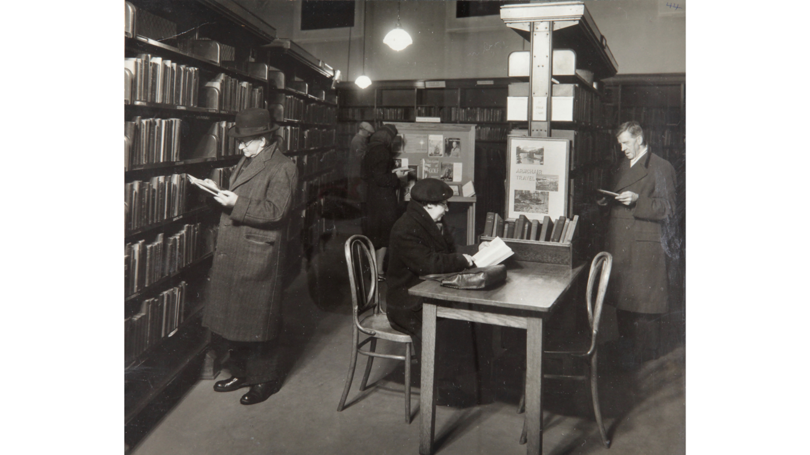 Black and white photograph of three patrons wearing heavy winter coats browsing books along tall wooden bookshelves inside a library aisle.