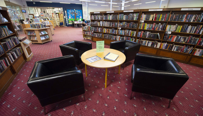 Seating area in Drumchapel Library. There are wooden bookshelves surrounding a table with 4 black leather chairs on a red carpet