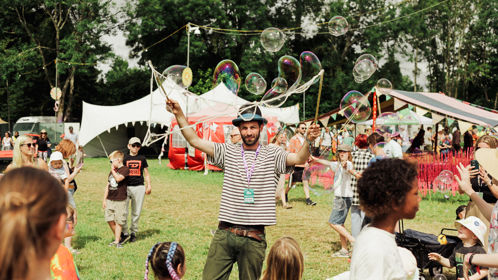 A children's entertainer creates large bubbles in a grassy area as children and adults look on