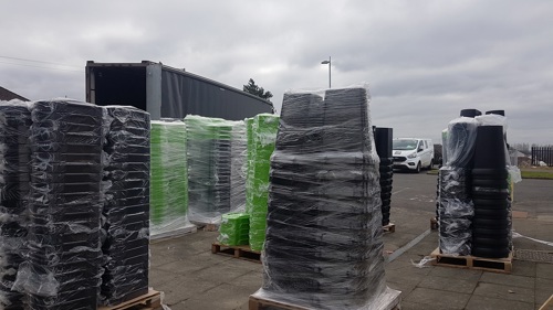 Several pallets of new bins in stacks, wrapped in plastic, on a tarmac area next to a large truck