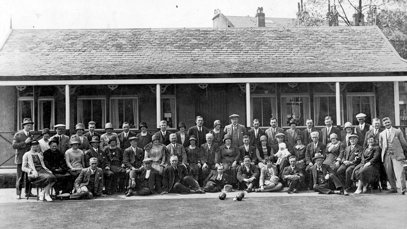 A black and white landscape photograph of a group of people wearing suits and dresses sitting outside a bowling club, the the lawn and bowls in front.