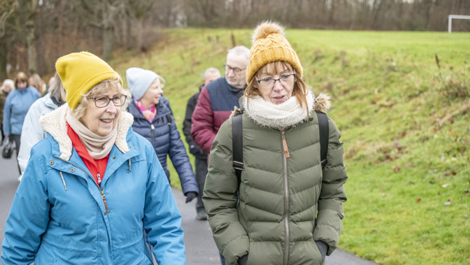 A group of older adults wearing warm puffer jackets and woolly hats walking outdoors on a grey day.