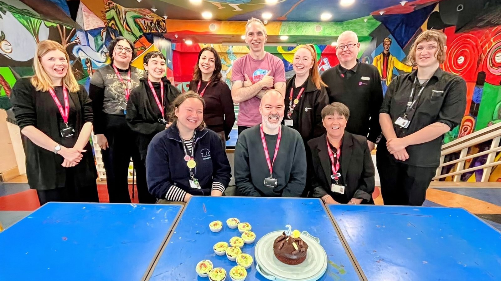 A group of twelve people stand together in a brightly painted room with colorful murals on the walls and ceiling. They wear name badges and smile toward the camera. In front of them are blue tables holding a chocolate cake and decorated cupcakes, suggesting a celebration. A staircase and vibrant artwork fill the background, creating a lively, creative atmosphere