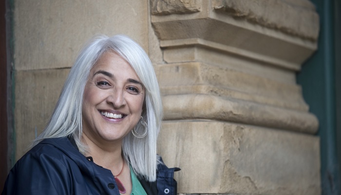 Portrait of a person with shoulder-length light-coloured hair standing beside a sandstone building. The person is wearing a dark jacket over a green top and looking towards the camera. Architectural stonework fills the background, with space to the right of the image.