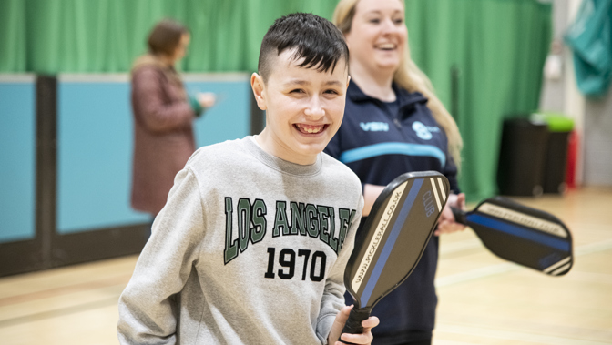 A smiling boy holding a pickleball paddle stands in a gymnasium with a woman behind him also holding a paddle, both appearing to enjoy the activity.