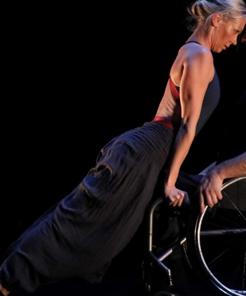 Two dancers, one female, one male and in a wheelchair, lean together, face to face in an intense pose