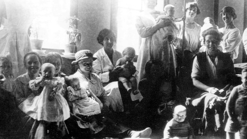 Black and white image showing a baby clinic circa 1900, mothers are holding babies and nurses are standing, there are large scales for weighing babies