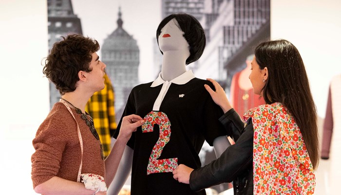 Two students adjusting a dress on a tailors model. The dress is black with a white collar in the style of Mary Quant.