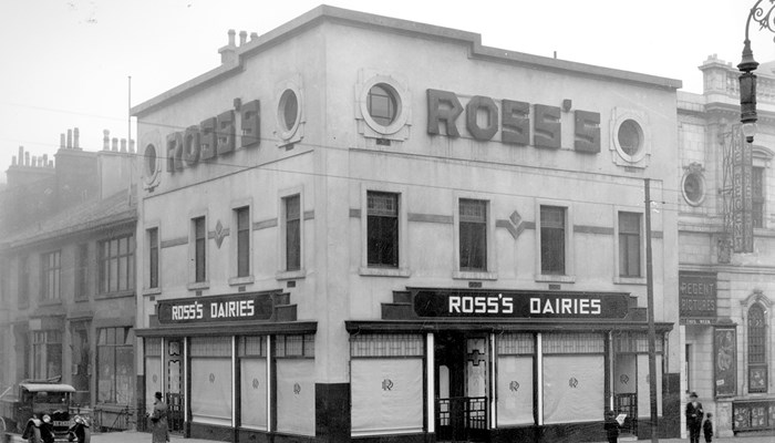 A black and white photo of Ross's Dairies three story shop front which sits on a corner of a busy road crossing, with people and cars passing by.