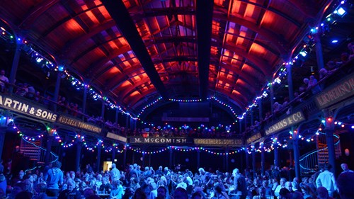 a gig setting indoors with blue lighting throughout. there are lots of people are fairy lights hanging from the balcony that surrounds the room