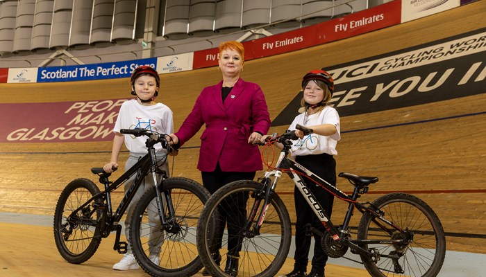 Child cyclists stand holding bicycles on wooden indoor velodrome either side of woman in purple jacket