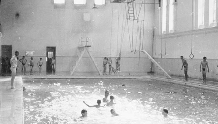 A black and white photo of lots of people having fun in a swimming pool. Some people are watching, some are swimming and others are about to jump in.