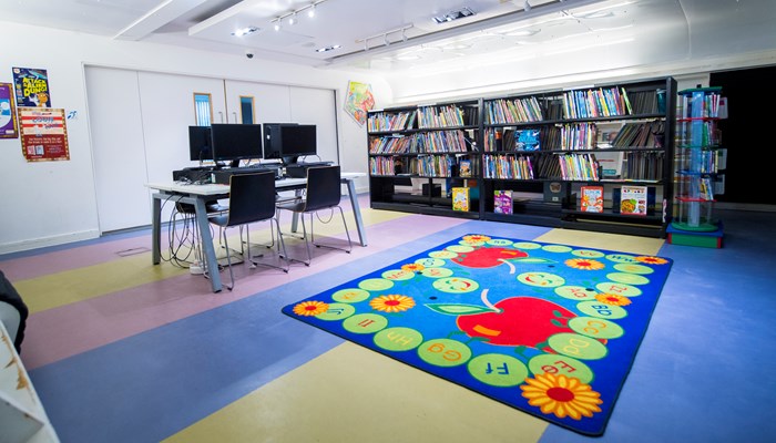 A spacious brightly lit area with 4 PC's in the middle of the room. Behind it there is a blue, green and orange rectangular children's rug on the floor