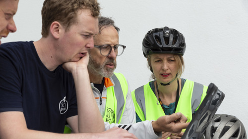 Three people look at an ipad with cycling helmets on.