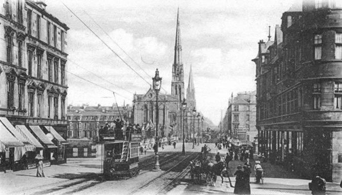 A black and white photo of a street with trams driving along the lines, tall tenement style shop buildings are on both sides with people walking on the pavement. You can also see the spires of a cathedral and a church in the background.