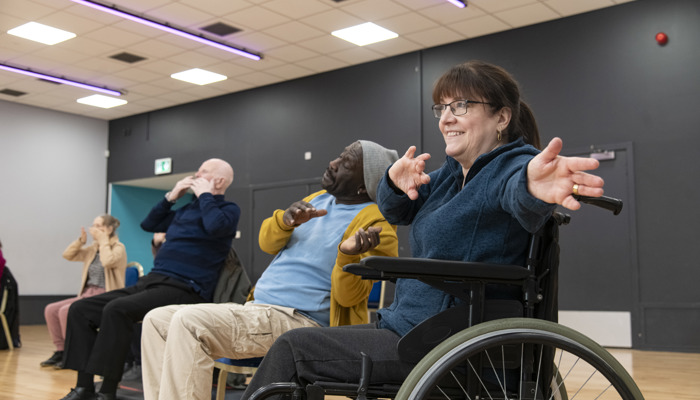 A group of people sitting down and waving their hands while taking part in an exercise class, one of them is in a wheelchair.