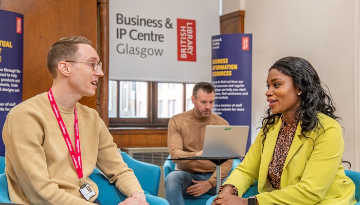 Two people are sitting talking to each other in the foreground as a third looks at a laptop screen in the background. A sign in the background reads Business and IP Centre Glasgow