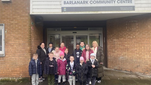 A group of 20 young people and adults wearing winter coats, standing outside a brick built community centre building
