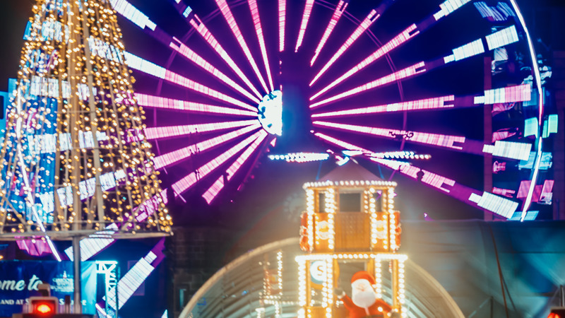Illuminated Ferris wheel and Christmas tree lights at a festive market, with a Santa display under a glass dome in the foreground.