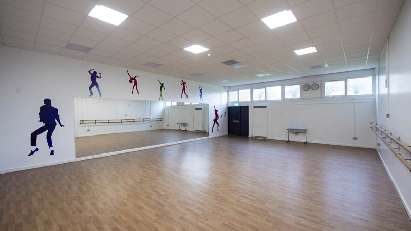 an empty dance studio with windows at the back of the room. the floor is wooden and the walls are white. there are five stickers on the wall of the outline of dancers and a bigger sticker of michael jackson dancing