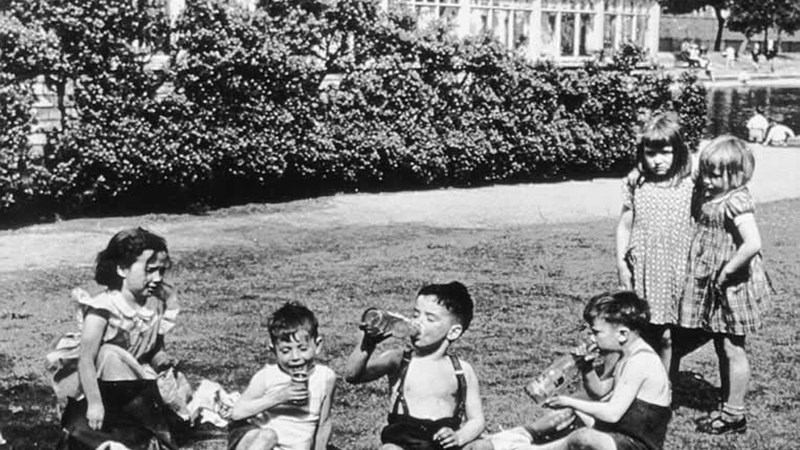 Black and white photo showing 6 children in a park having a picnic, three of the children are boys and are drinking from bottles of juice.
